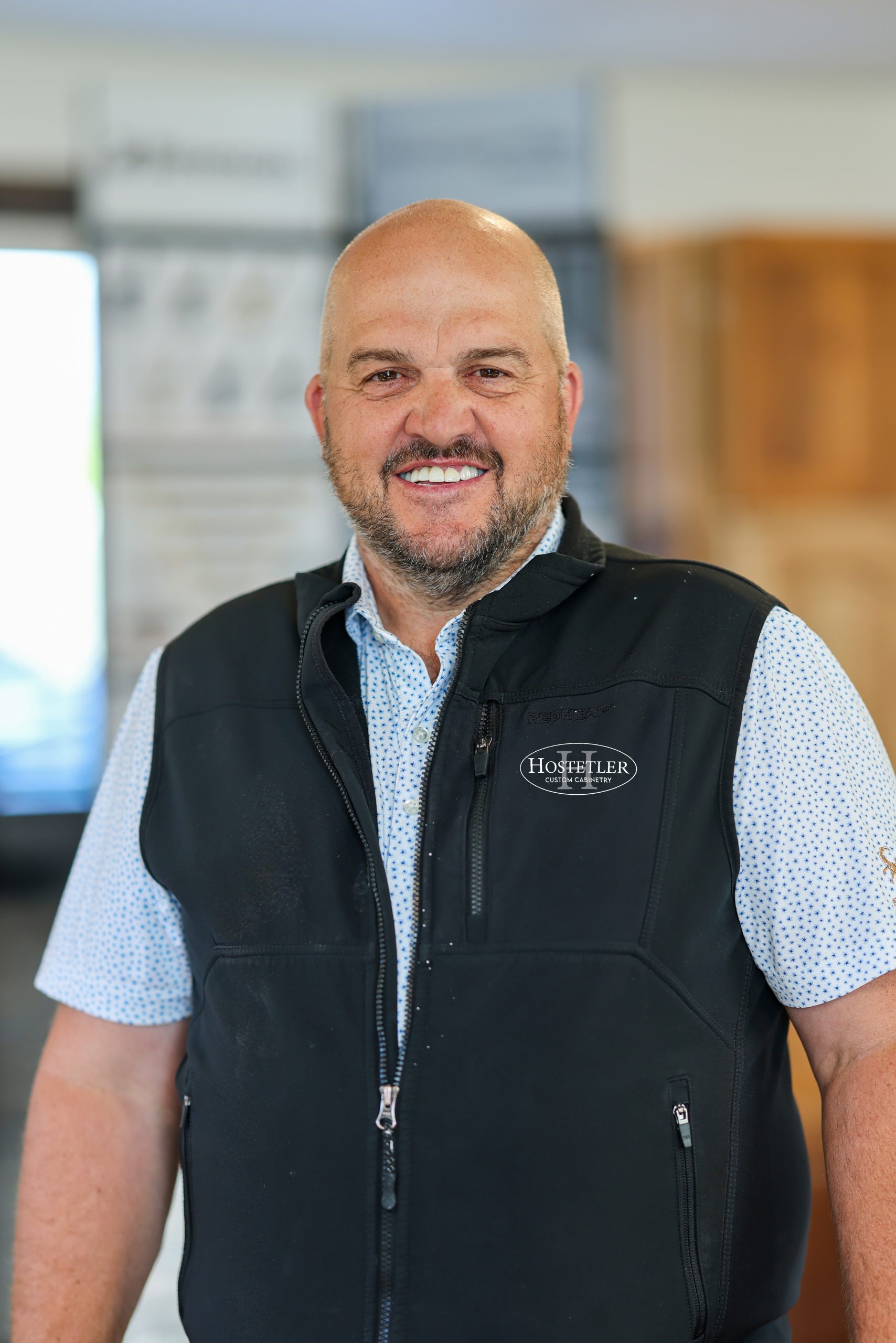 Friendly team member wearing a black Hostetler logo vest stands in a welcoming, well-lit space, ready to help guide you through your custom cabinetry journey.