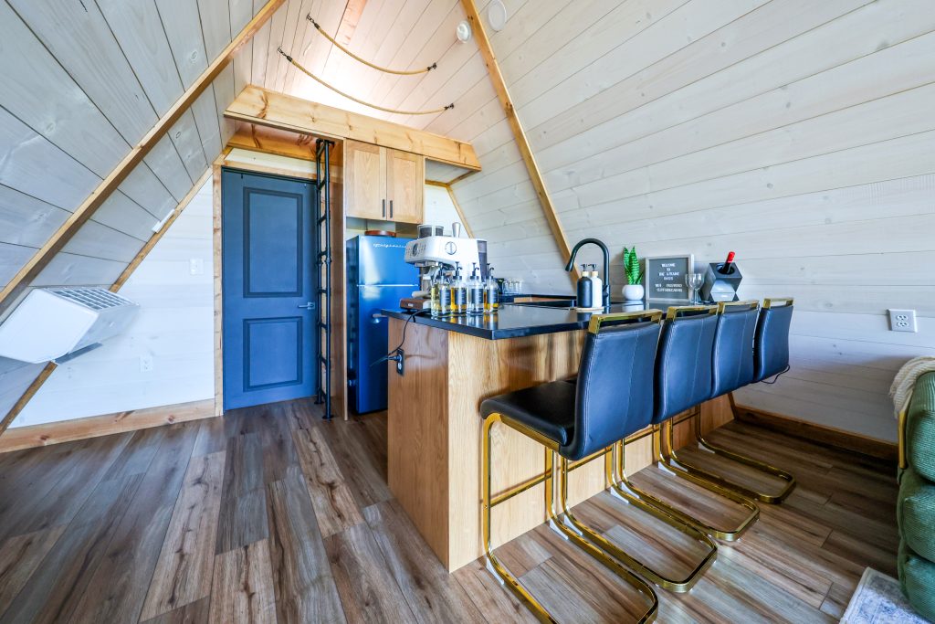 Inviting A-frame cabin kitchen showcasing thoughtfully crafted Hostetler custom wood cabinets, a cheerful blue fridge, welcoming bar counter, and four sleek black barstools—designed to guide you toward the perfect gathering space even if you’re unsure where to start.