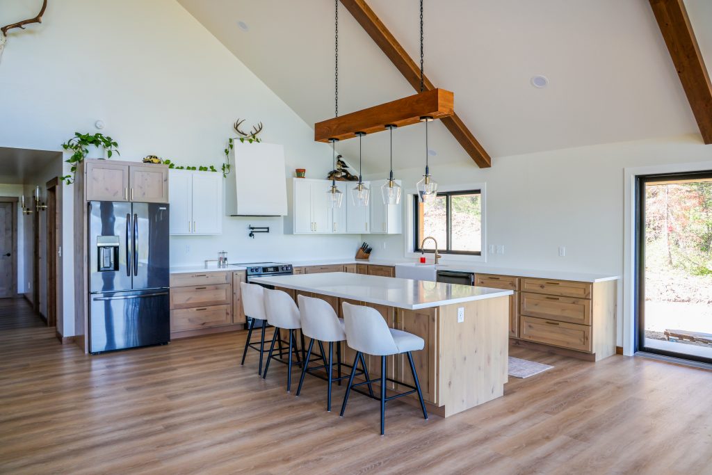 Inviting modern kitchen featuring warm wood floors and exposed beams, highlighted by light wood Hostetler custom cabinets designed for both beauty and everyday living. A spacious island with four white chairs offers a welcoming space to gather, while sleek stainless steel appliances, elegant pendant lights, and greenery on top of the cabinets add comfort and style. Expansive window and glass door fill the space with natural light—reflecting a thoughtfully crafted environment ready to evolve with your needs.