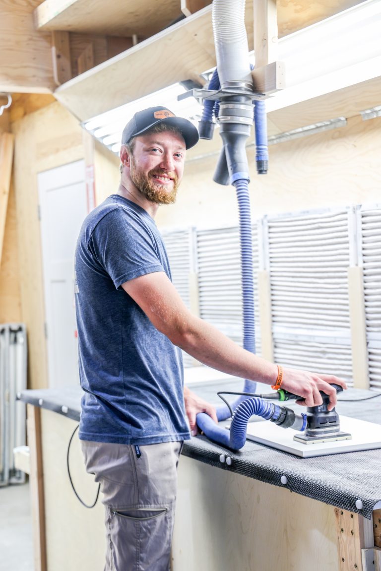 Hostetler Custom Cabinets shop employee smiling while sanding cabinet components.