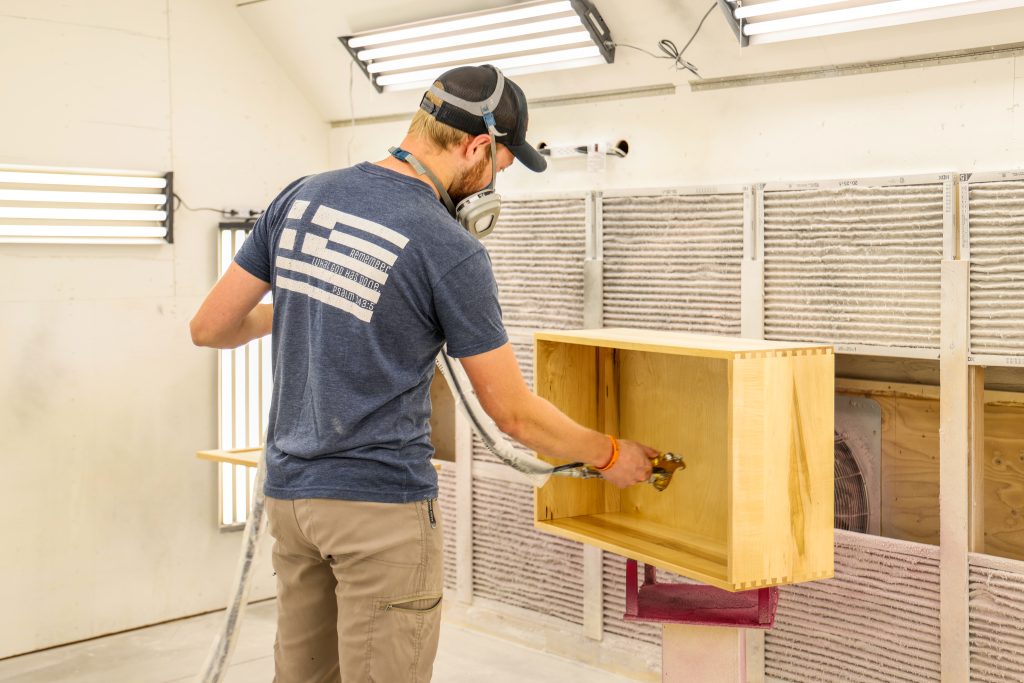 In our bright workshop, a skilled craftsman in a safety mask carefully applies finish to custom wooden cabinet drawers, guiding each step of the process with the attention and support that defines Hostetler Custom Cabinetry.